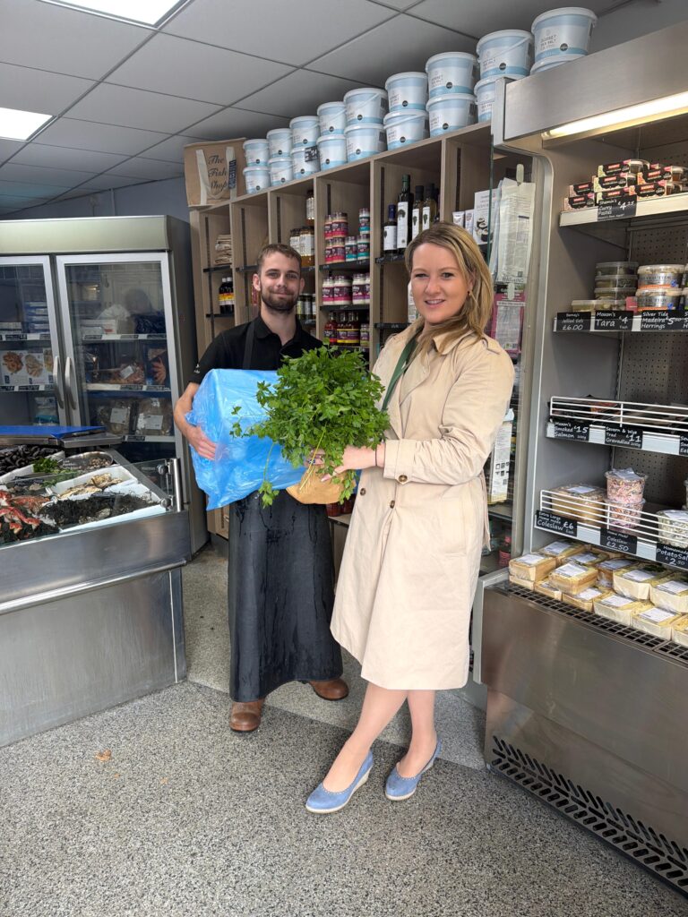A woman in a beige trench coat holds fresh herbs next to a man in a dark apron holding a large box of Megrim wrapped in blue. They're inside a store with shelves and a seafood display, smiling warmly.