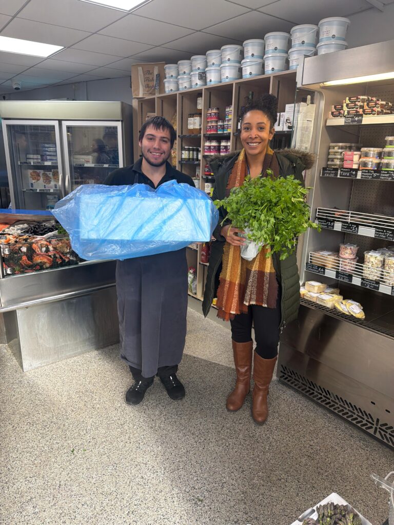 A woman in a long green coat holds fresh herbs next to a man in a dark apron holding a large box of Megrim wrapped in blue. They're inside a store with shelves and a seafood display, smiling warmly.